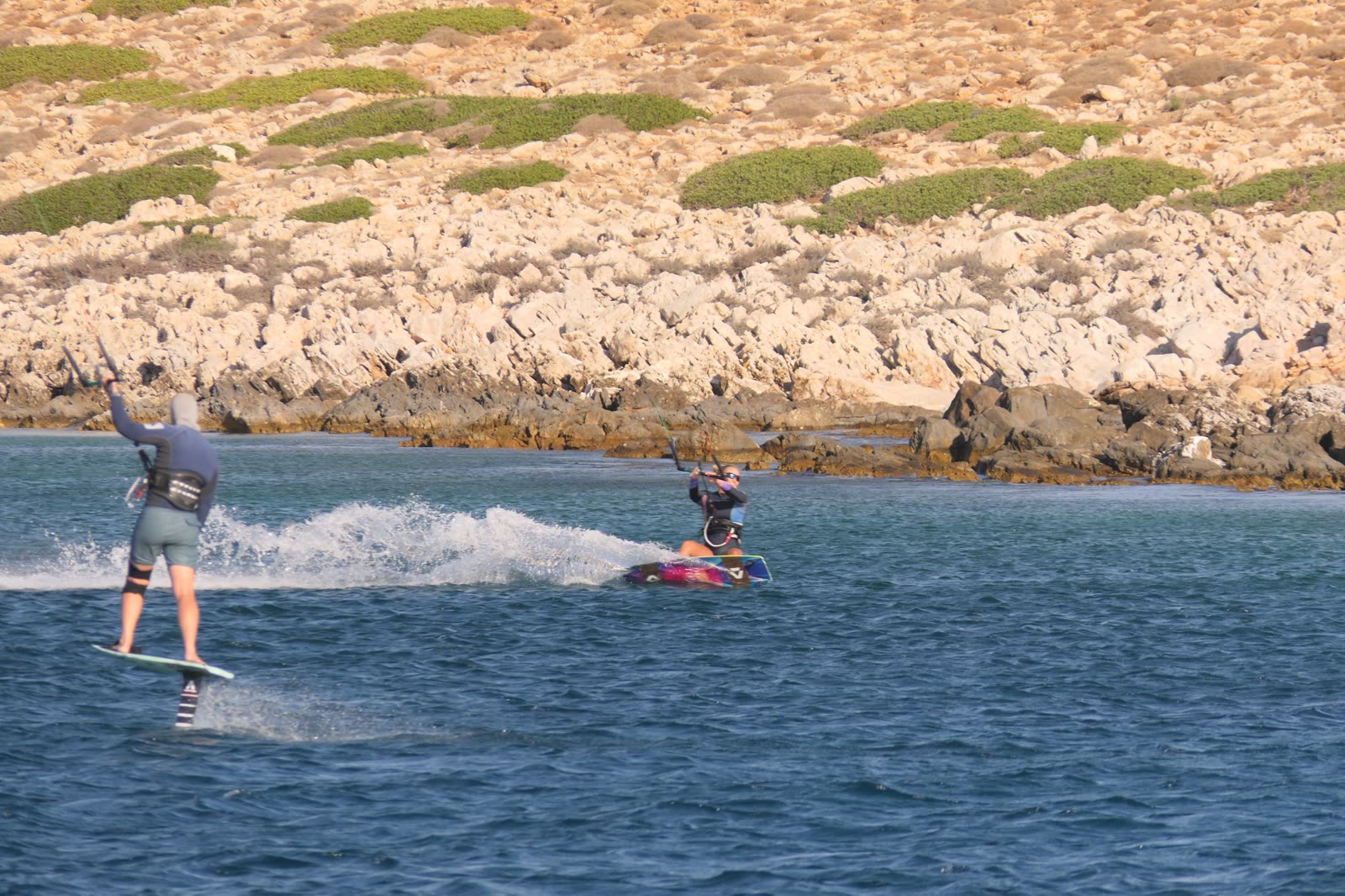 A hydrofoil rider and a kiteboarder sharing the water beside the rocky coast