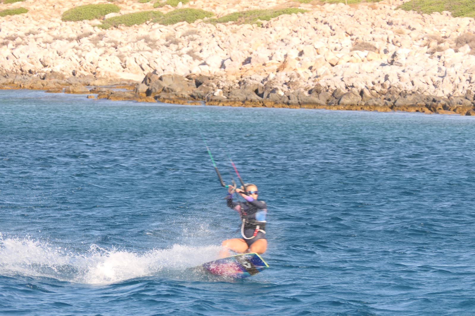 Tina kitesurfing across blue water beside the rocky coast in Greece